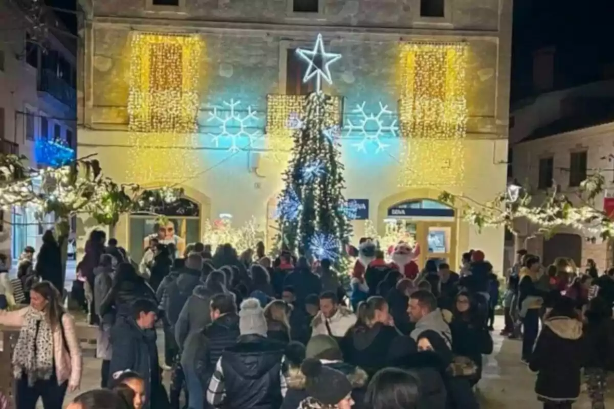 Una multitud de personas se reúne en una plaza decorada con luces navideñas y un árbol iluminado frente a un edificio adornado con estrellas y copos de nieve.