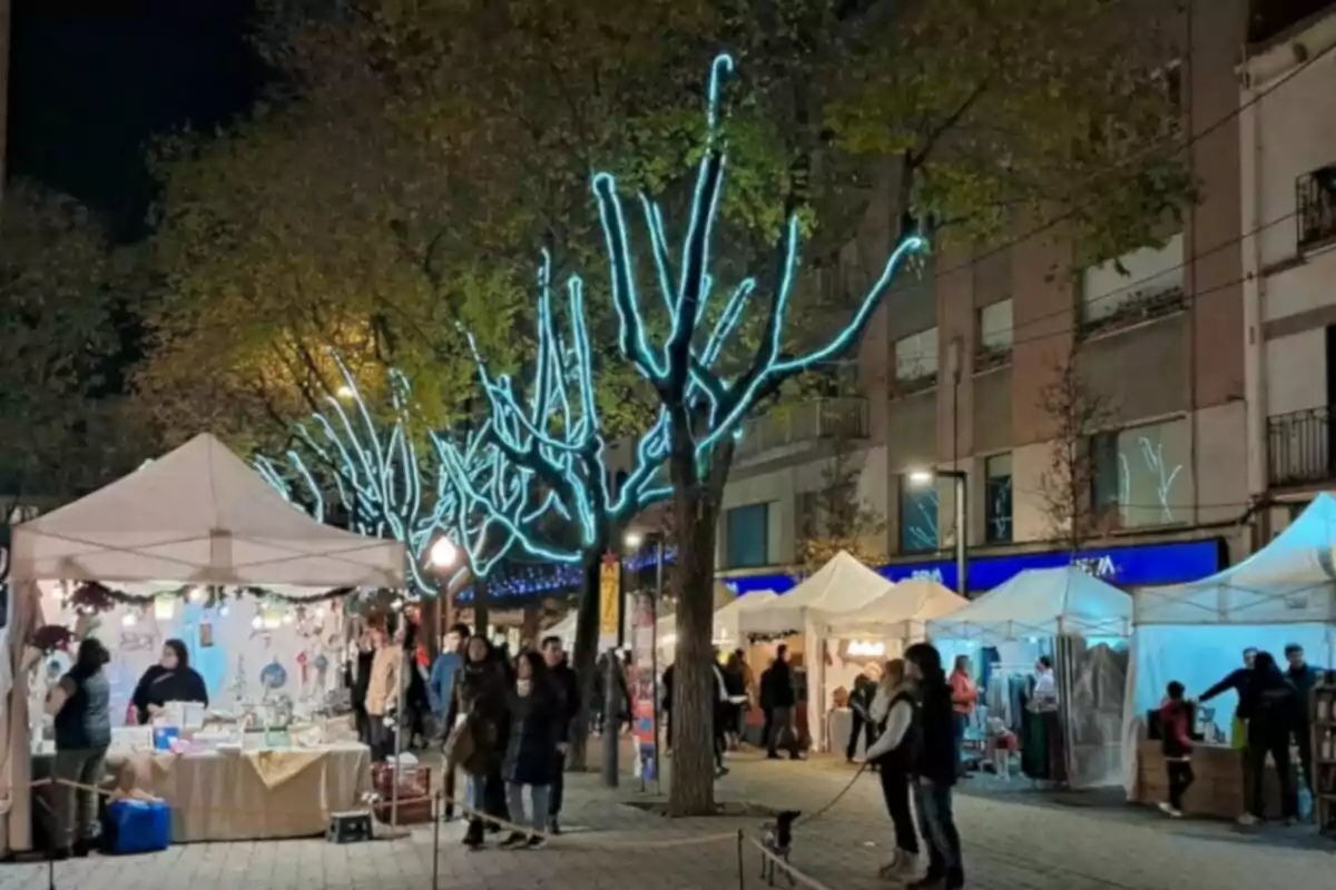 Una feria nocturna con puestos de venta iluminados y árboles decorados con luces azules.