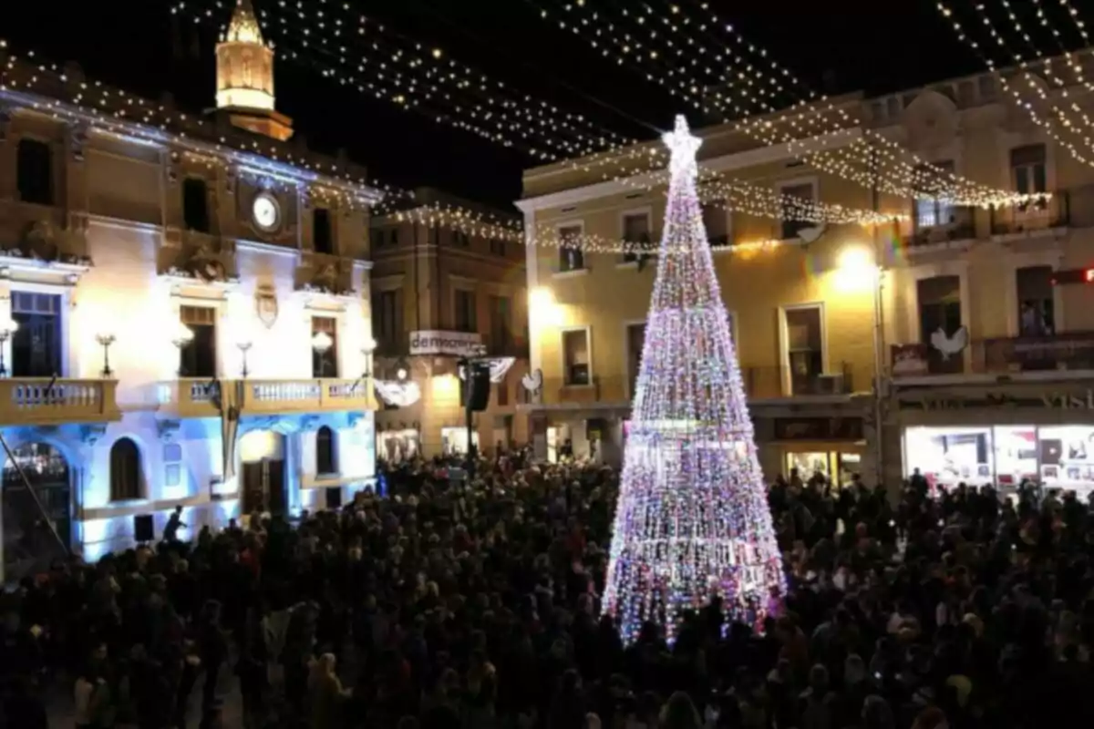 Una plaza iluminada con un gran árbol de Navidad decorado con luces brillantes rodeado de una multitud de personas bajo un cielo nocturno adornado con guirnaldas de luces.