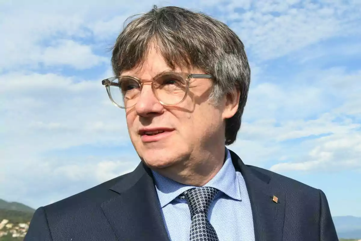 Carles Puigdemont con gafas y traje oscuro mirando hacia la derecha con un cielo azul y nubes de fondo.