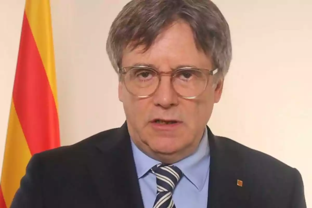 Un hombre con gafas y traje oscuro frente a una bandera con franjas rojas y amarillas.