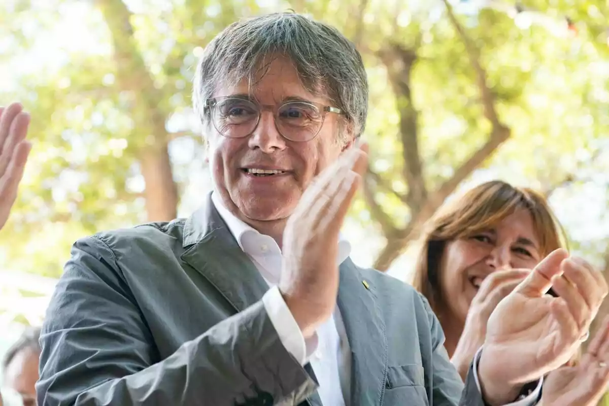 Carles Puigdemont aplaudiendo al aire libre con Laura Borràs sonriendo en el fondo.