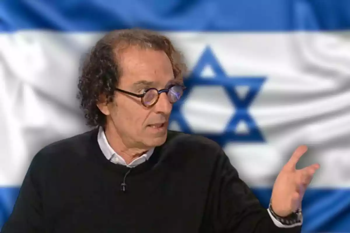 Un hombre con gafas redondas y cabello rizado gesticula mientras habla, con una bandera de Israel de fondo.