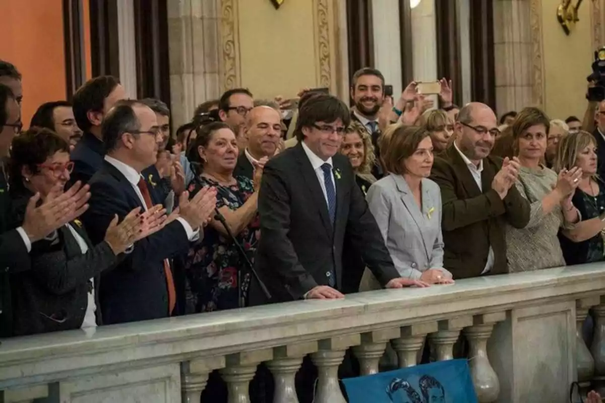 Un grupo de personas aplaude en un evento formal en un edificio con columnas y decoración clásica.