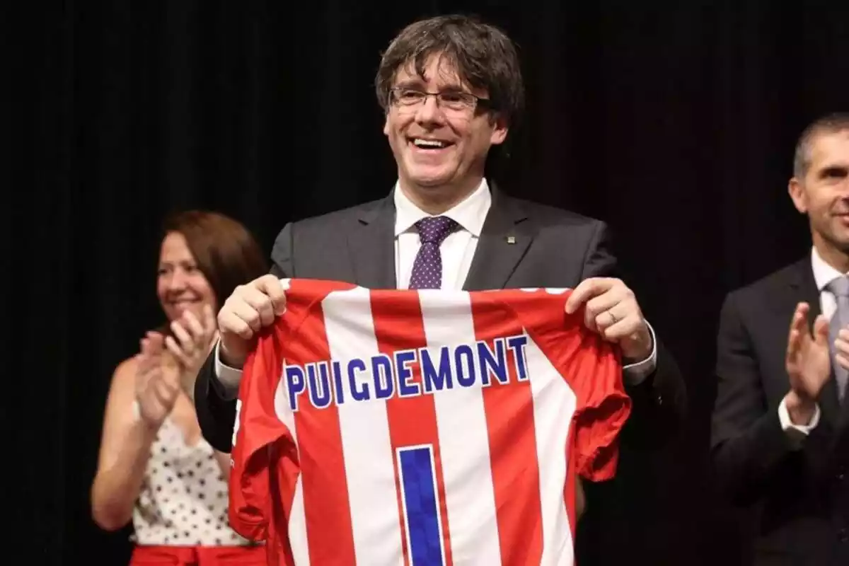Plano medio de Carles Puigdemont sonriendo y mostrando una camiseta del Girona FC con el nombre de Puigdemont y el dorsal número 1