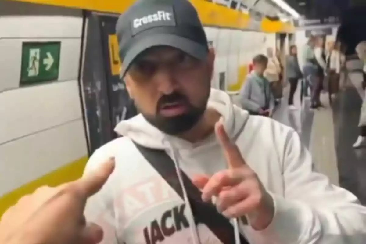 Un hombre con gorra y sudadera blanca está en una estación de metro, señalando con el dedo mientras otra persona lo señala a él.