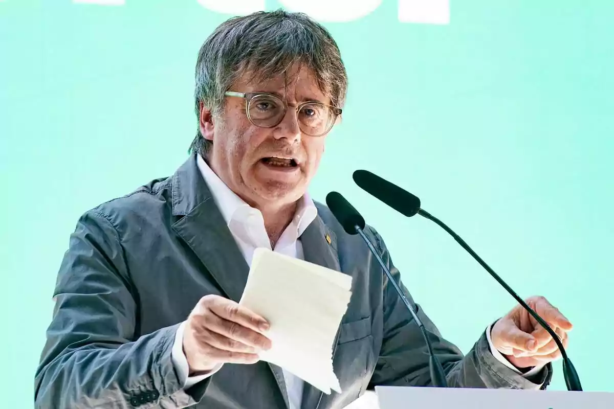 Carles Puigdemont hablando en un podio con micrófonos, sosteniendo papeles en la mano, fondo verde.