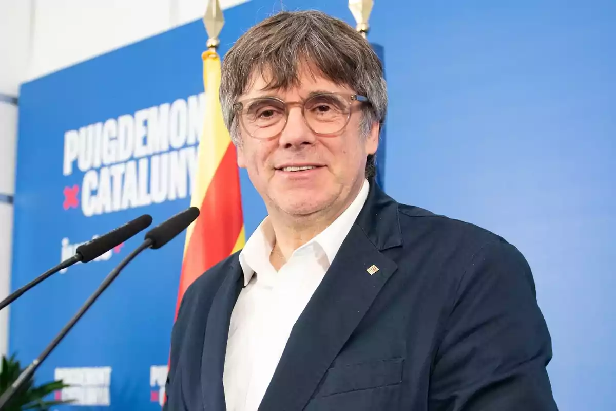 Hombre con gafas y cabello canoso en una conferencia de prensa frente a un fondo azul con texto y una bandera catalana.