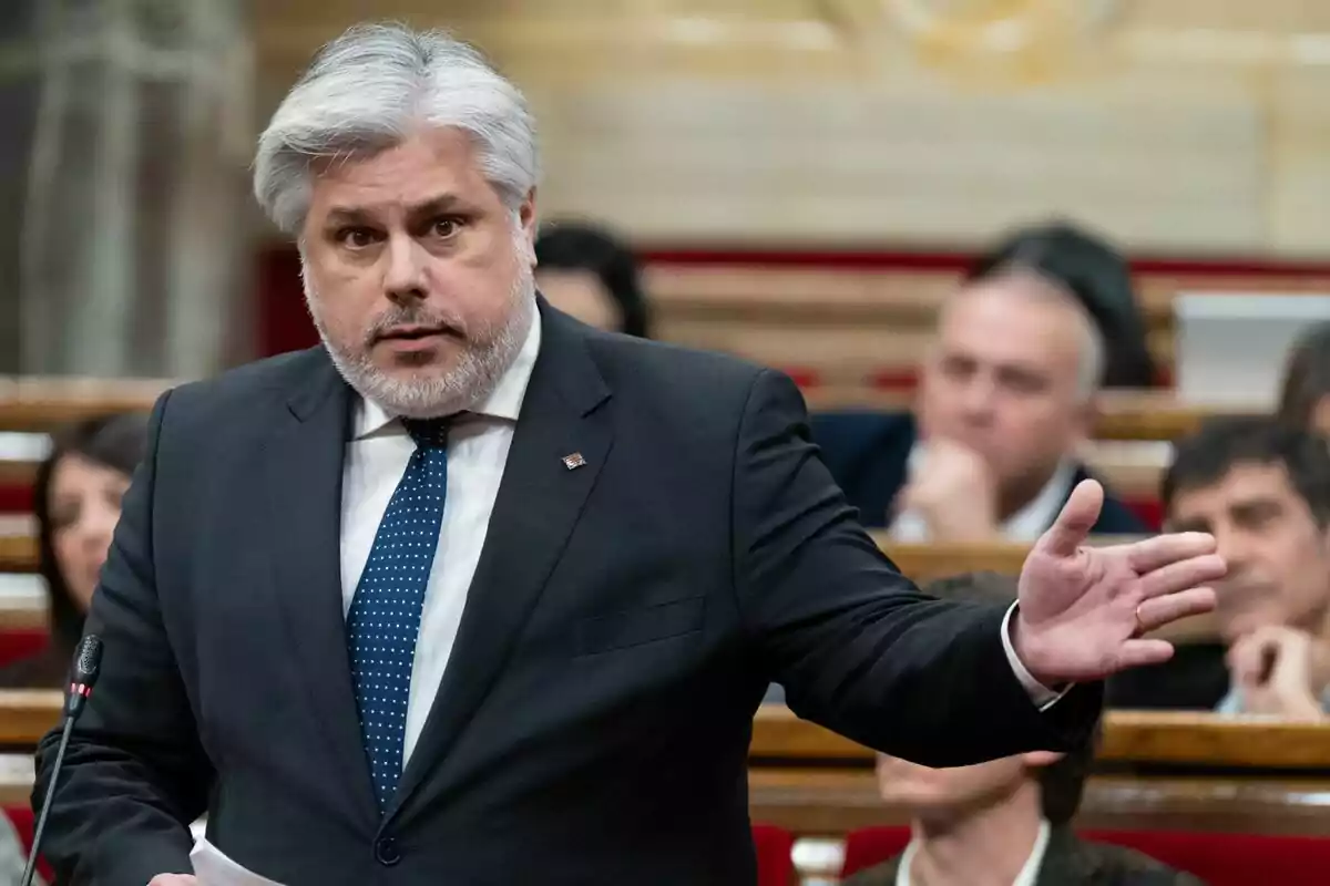 Un hombre de cabello canoso y barba, vestido con traje y corbata, hablando en un podio con un micrófono, con personas sentadas al fondo.