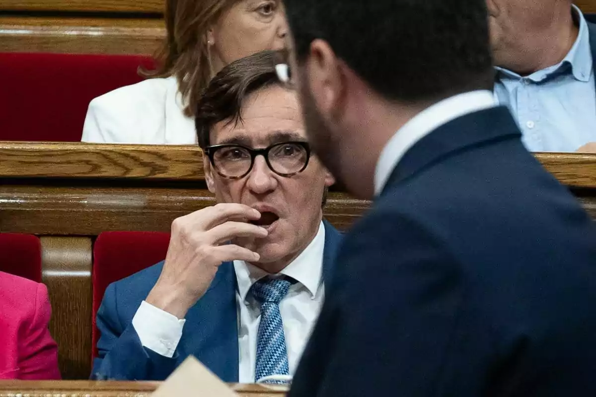 Un hombre con gafas y traje azul está sentado en un banco de madera, llevándose la mano a la boca mientras observa a otra persona de espaldas.