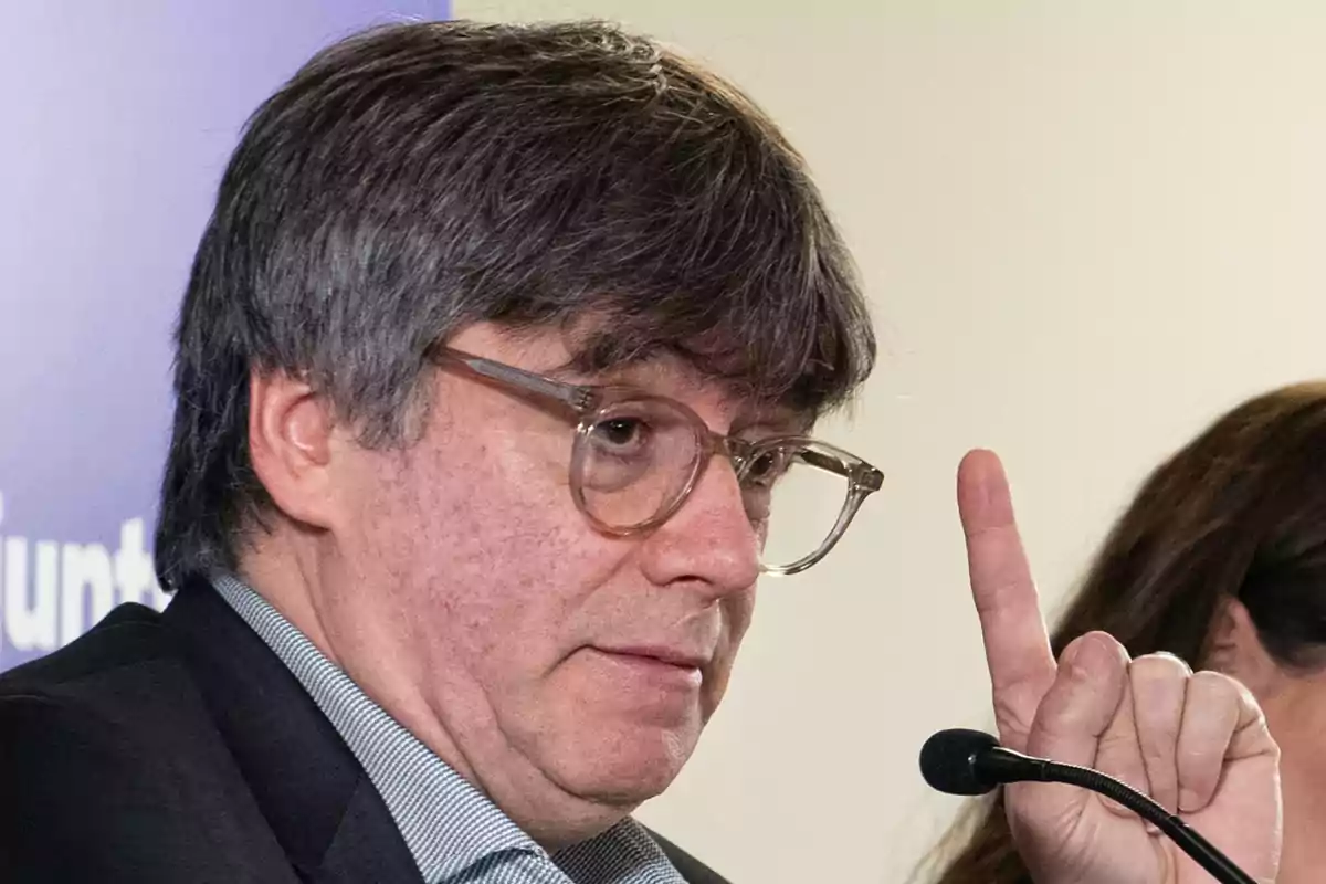 Hombre con gafas y cabello canoso levantando un dedo mientras habla en un micrófono.