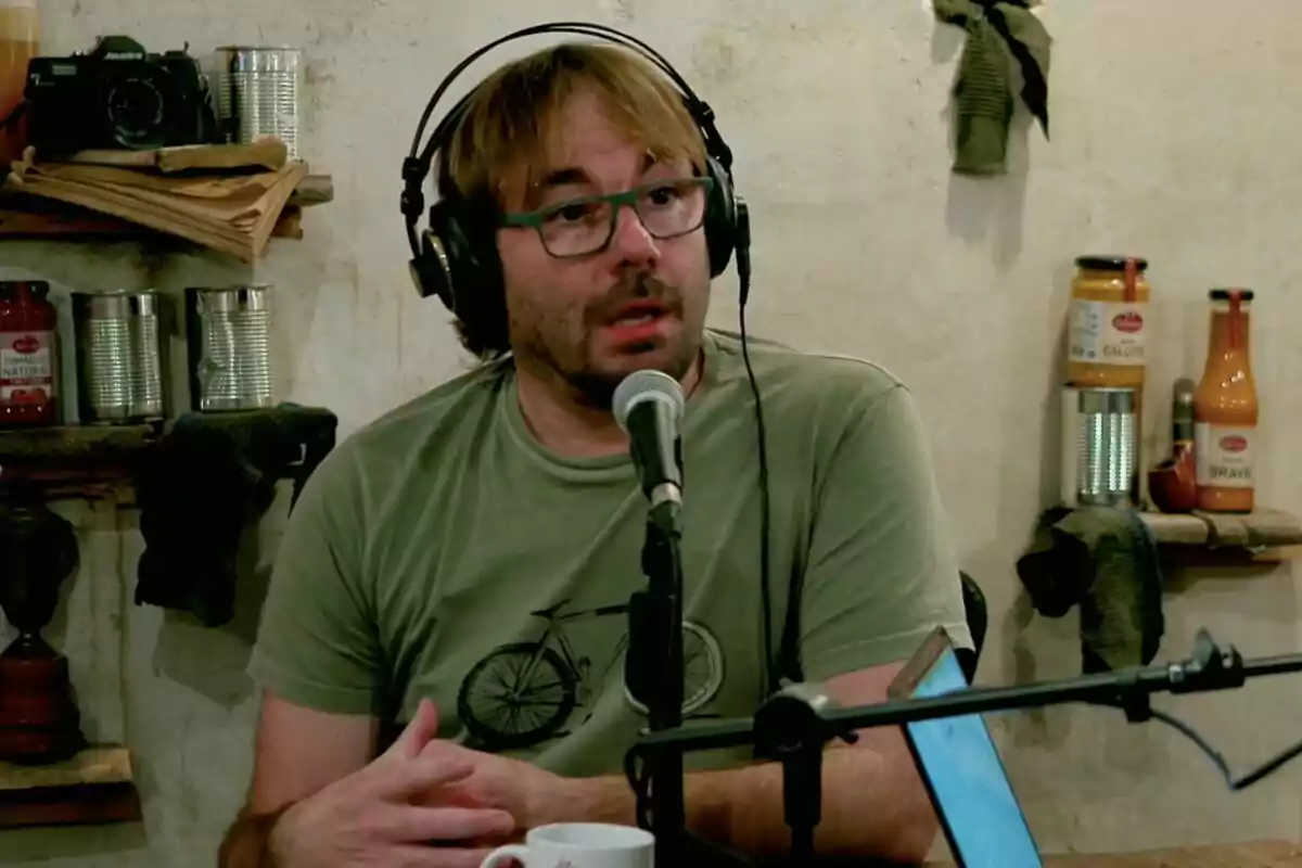 Quim Masferrer con gafas y auriculares habla frente a un micrófono en un entorno que parece ser un estudio de grabación con estantes llenos de latas y botellas en el fondo. Quim Masferrer con gafas y auriculares habla frente a un micrófono en un entorno que parece ser un estudio de grabación con estantes llenos de latas y botellas en el fondo.