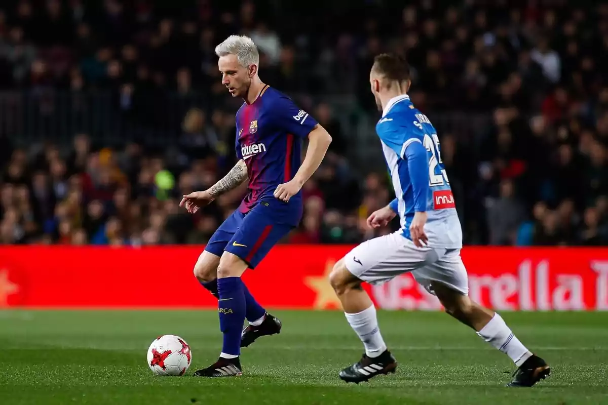 Un jugador del Barcelona controla el balón mientras un rival del Espanyol lo marca durante un partido de fútbol en un estadio lleno de espectadores. Un jugador del Barcelona controla el balón mientras un rival del Espanyol lo marca durante un partido de fútbol en un estadio lleno de espectadores.