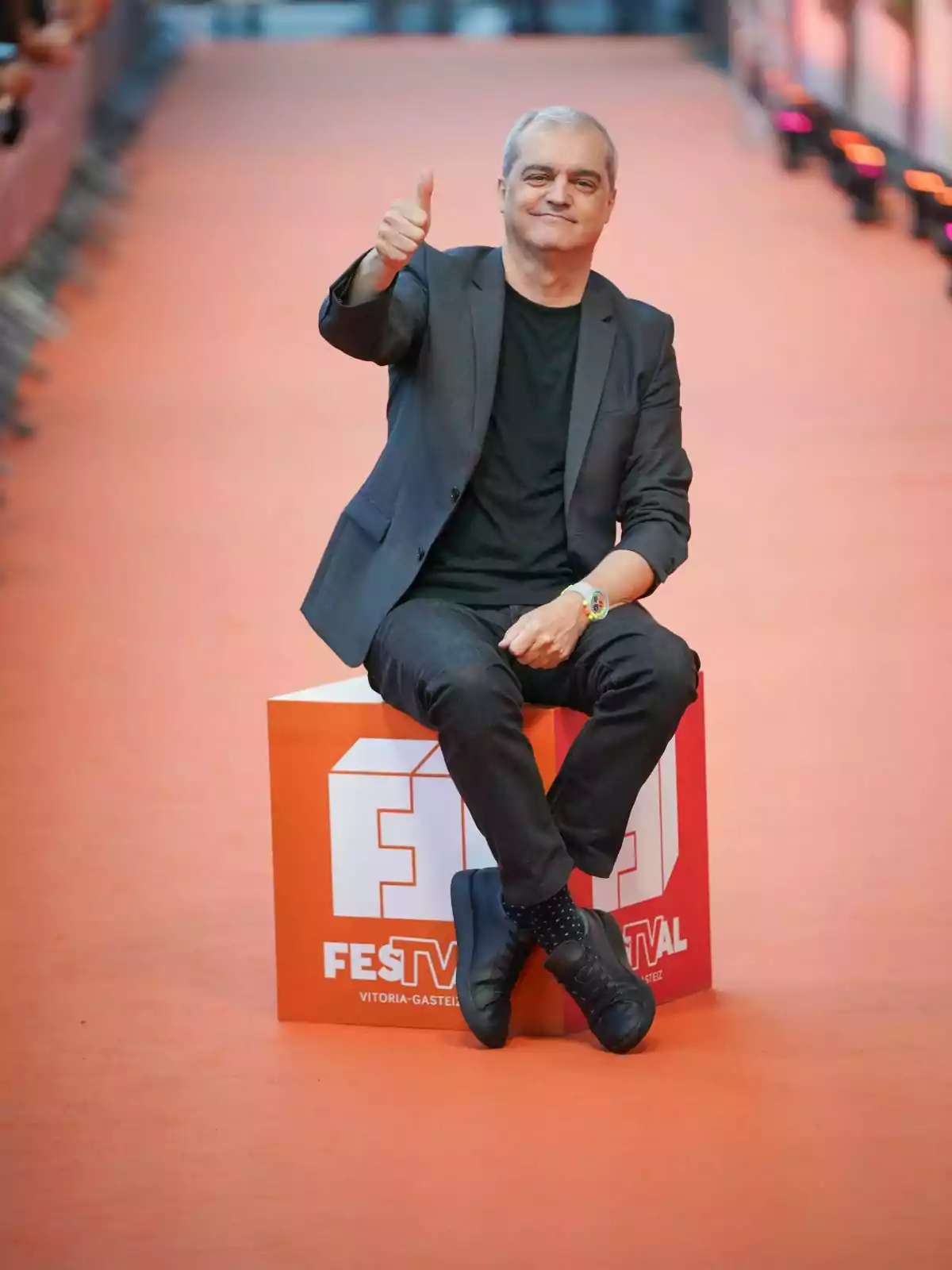 Ramón García sentado en un cubo naranja con el logo de FesTVal, haciendo un gesto de aprobación con el pulgar hacia arriba, en una alfombra roja. Ramón García sentado en un cubo naranja con el logo de FesTVal, haciendo un gesto de aprobación con el pulgar hacia arriba, en una alfombra roja.