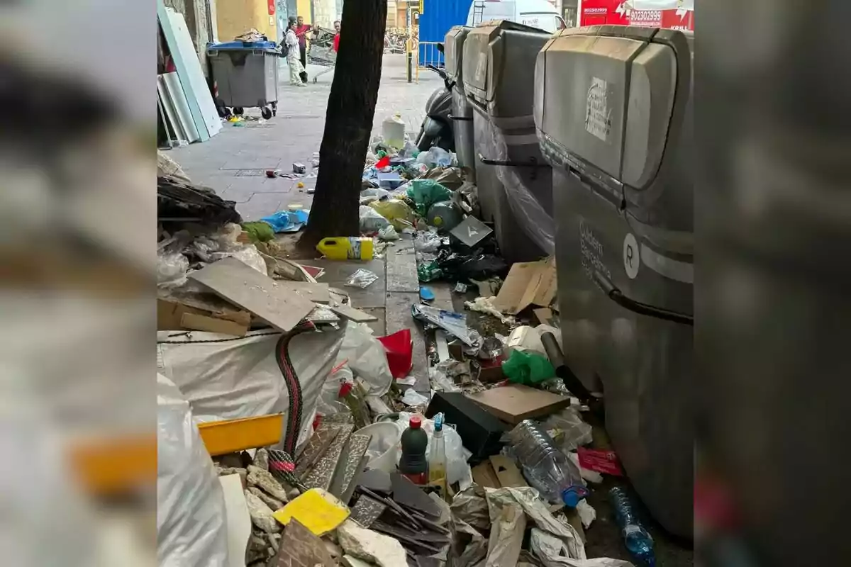 Una acera llena de basura y escombros junto a contenedores de reciclaje.