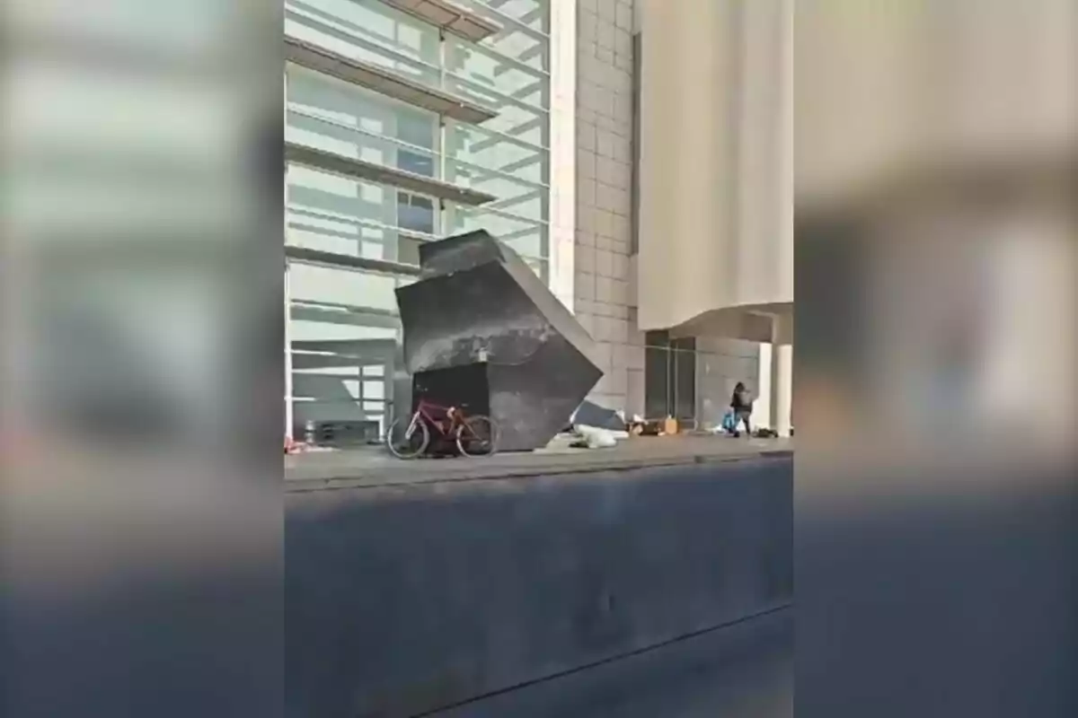 Una bicicleta roja está estacionada junto a una escultura geométrica negra frente a un edificio moderno con paredes de vidrio y concreto.