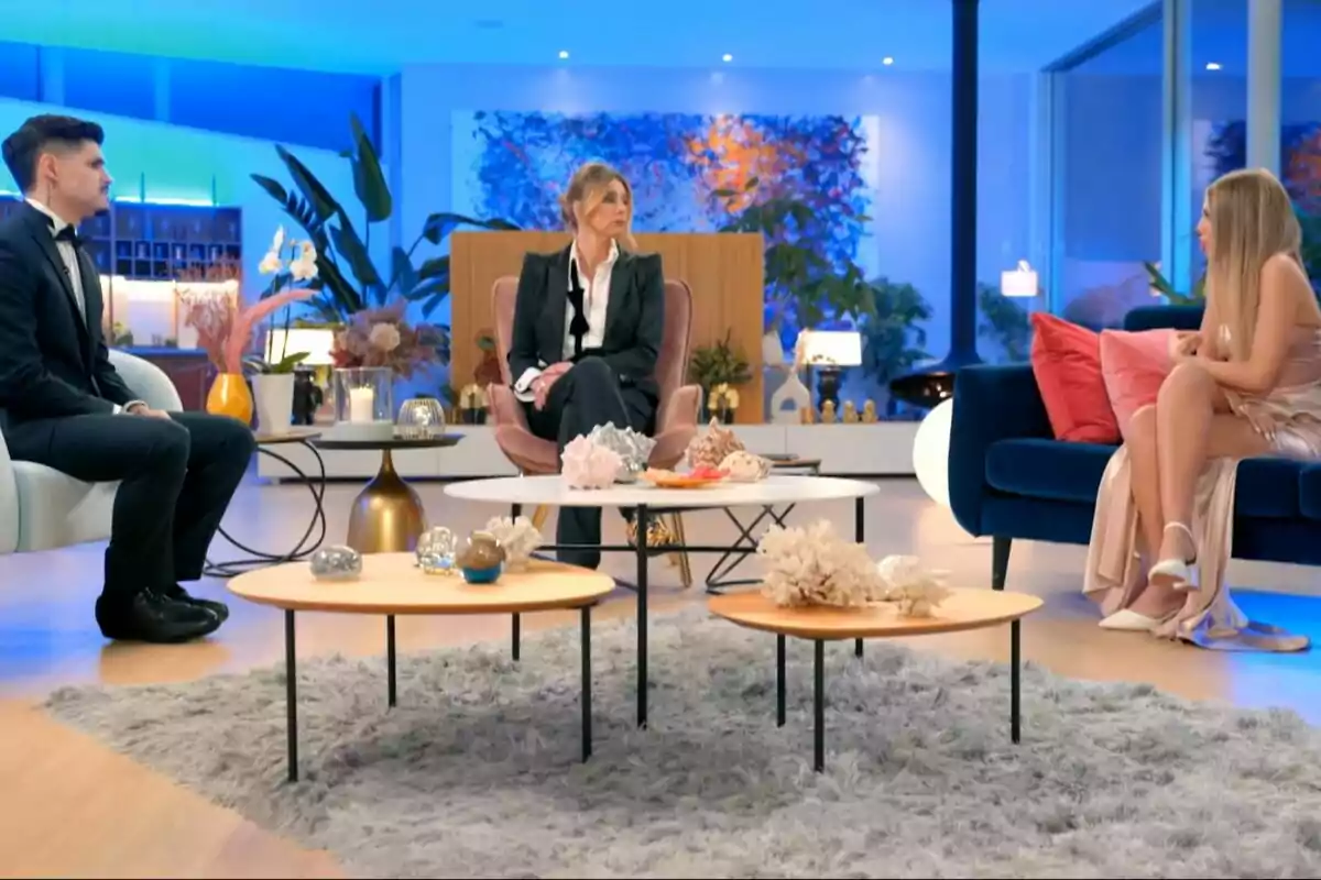 Álvaro, Sandra Barneda y Alba sentadas en un moderno salón con decoración elegante y luces azules, conversando alrededor de mesas de centro con adornos en La Isla de las Tentaciones. Álvaro, Sandra Barneda y Alba sentadas en un moderno salón con decoración elegante y luces azules, conversando alrededor de mesas de centro con adornos en La Isla de las Tentaciones.