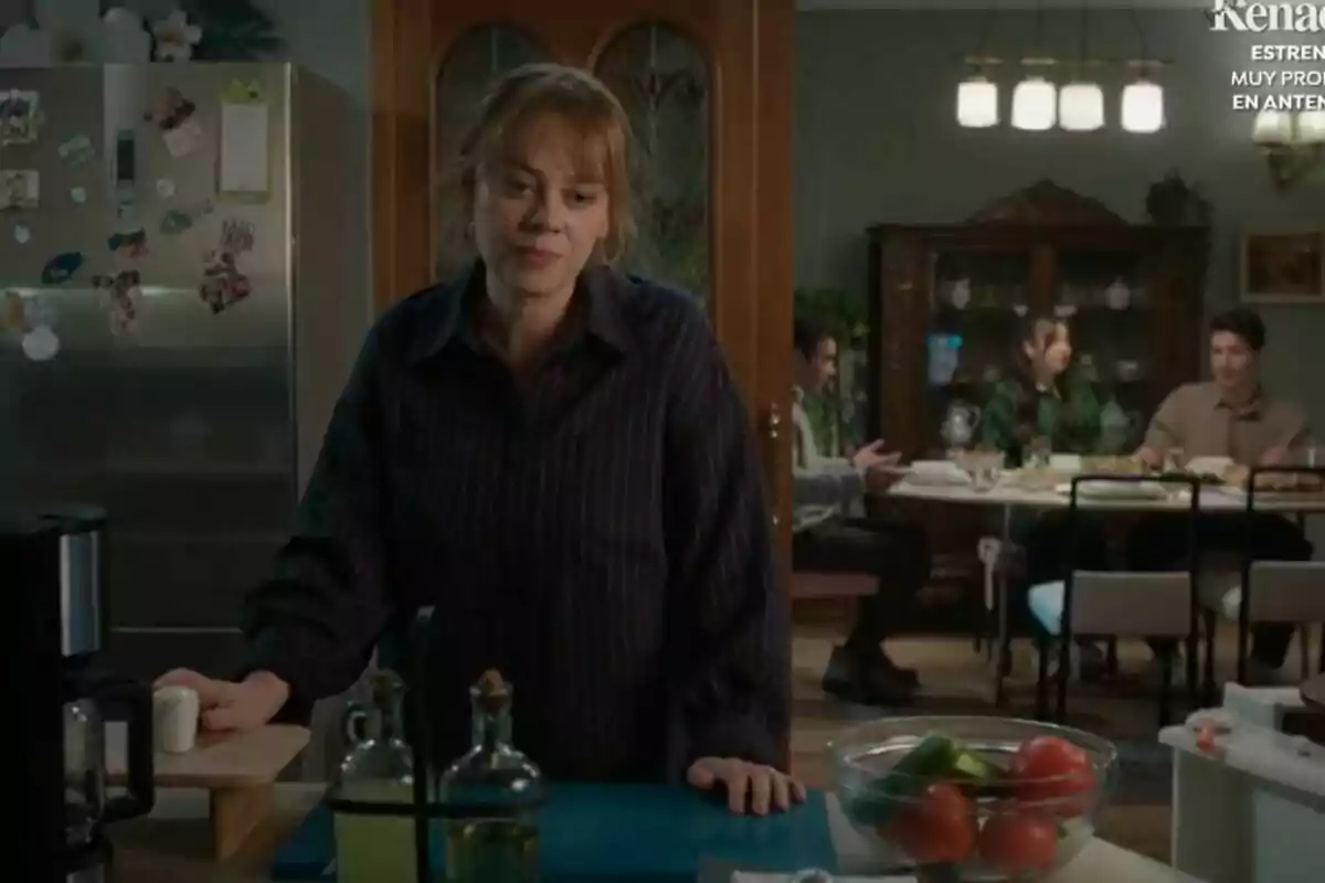 Una mujer está en una cocina con una expresión pensativa mientras en el fondo se ve a varias personas sentadas alrededor de una mesa en un comedor iluminado, en la nueva serie turca Renacer de Antena 3.