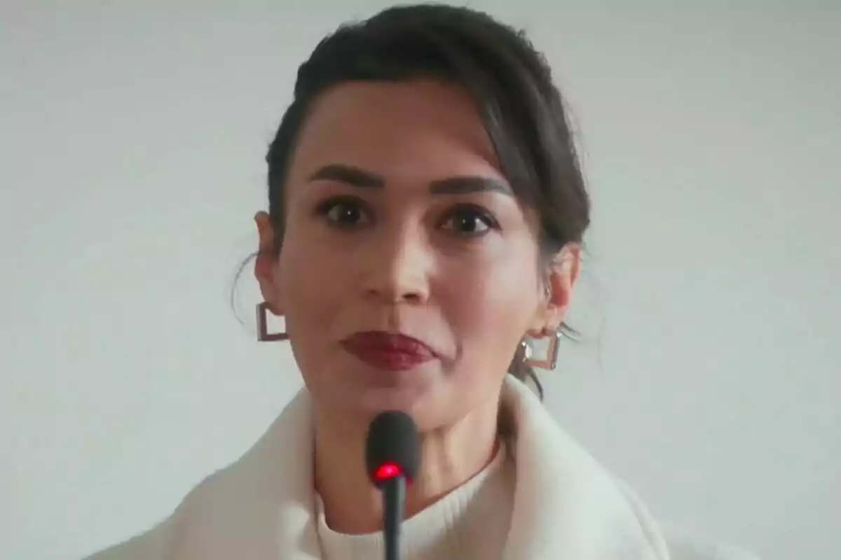 Rengin de cabello oscuro y labios rojos hablando frente a un micrófono con fondo claro en Renacer Rengin de cabello oscuro y labios rojos hablando frente a un micrófono con fondo claro en Renacer
