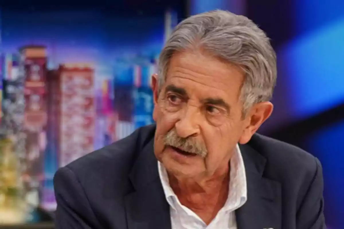 Miguel Ángel Revilla con bigote y cabello canoso, vestido con traje oscuro y camisa blanca, en un entorno de fondo colorido y desenfocado en 'El Hormiguero'.