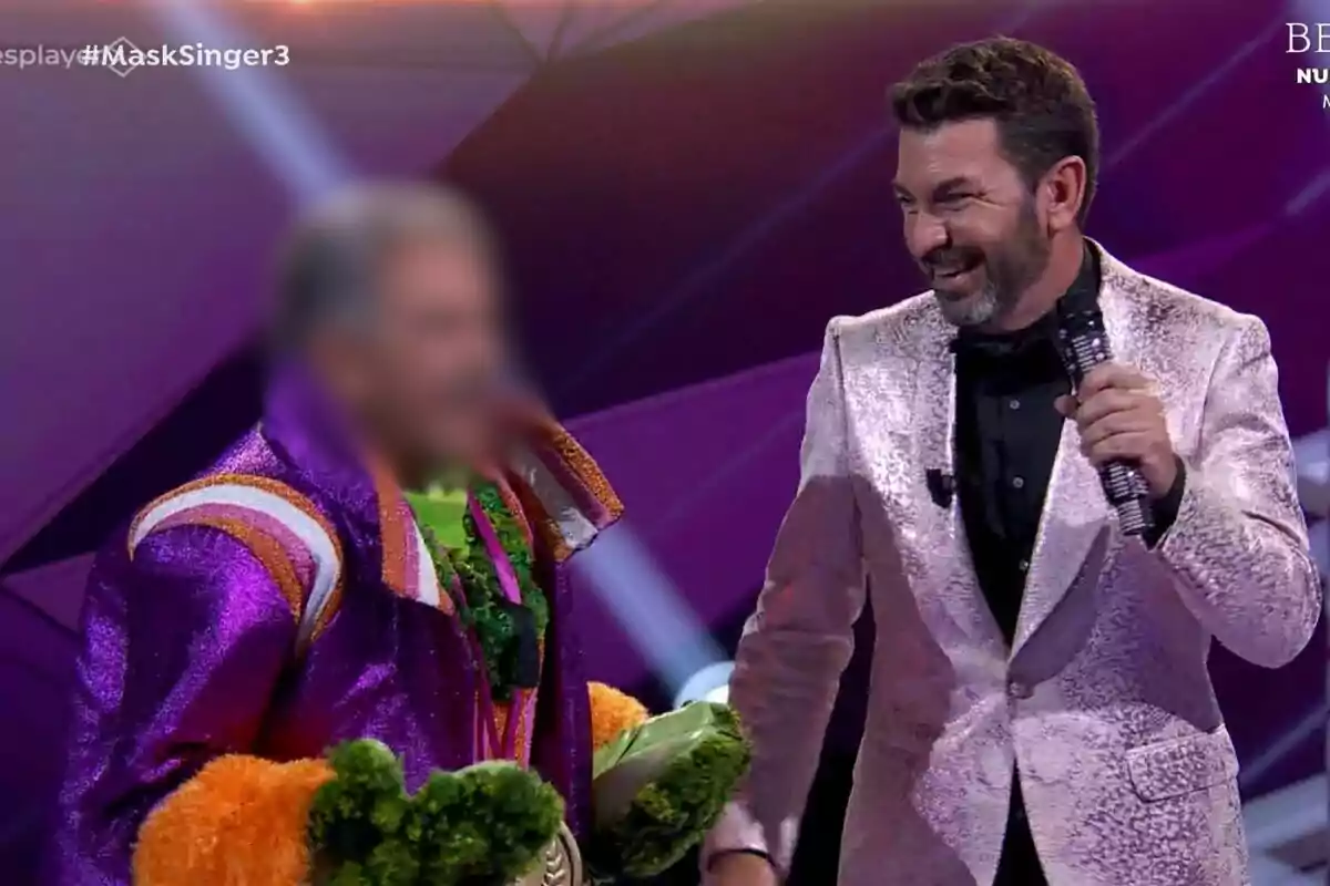 Arturo Valls sonriente sostiene un micrófono mientras interactúa con una persona disfrazada en un escenario colorido en Mask Singer.