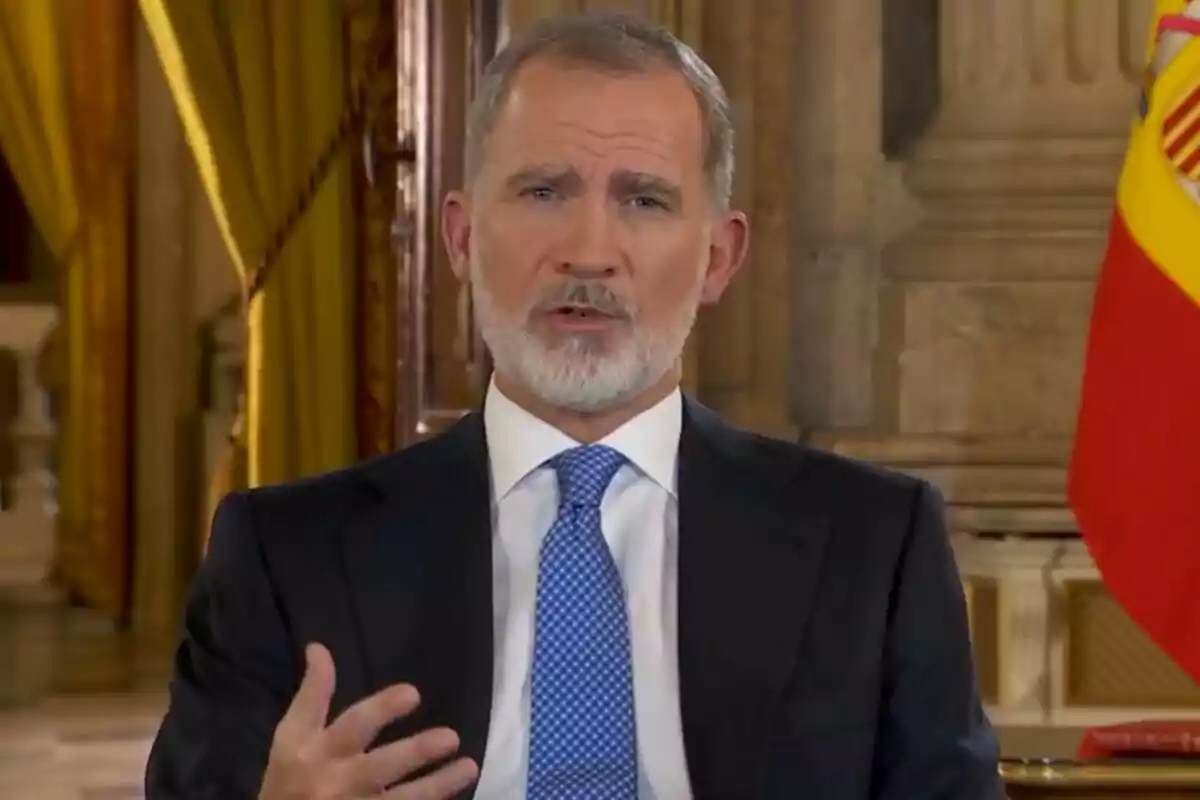 Un hombre con barba y traje oscuro habla frente a una bandera en un entorno elegante.