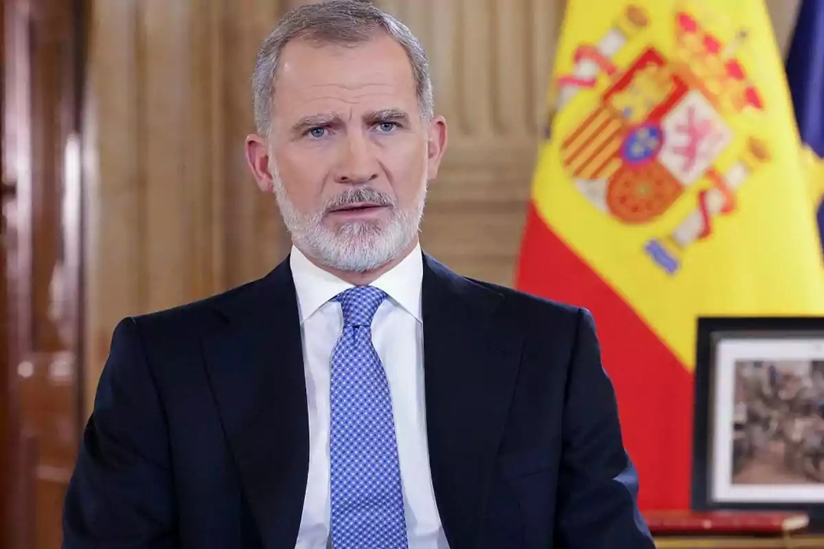 Felipe VI con barba y traje oscuro está frente a una bandera de España.