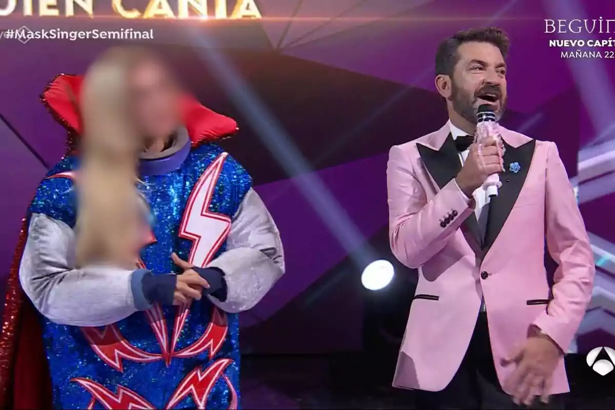 Arturo Valls con un traje rosa sostiene un micrófono mientras está de pie junto a una persona disfrazada con un traje azul brillante y detalles de rayos en un escenario iluminado en Mask Singer.