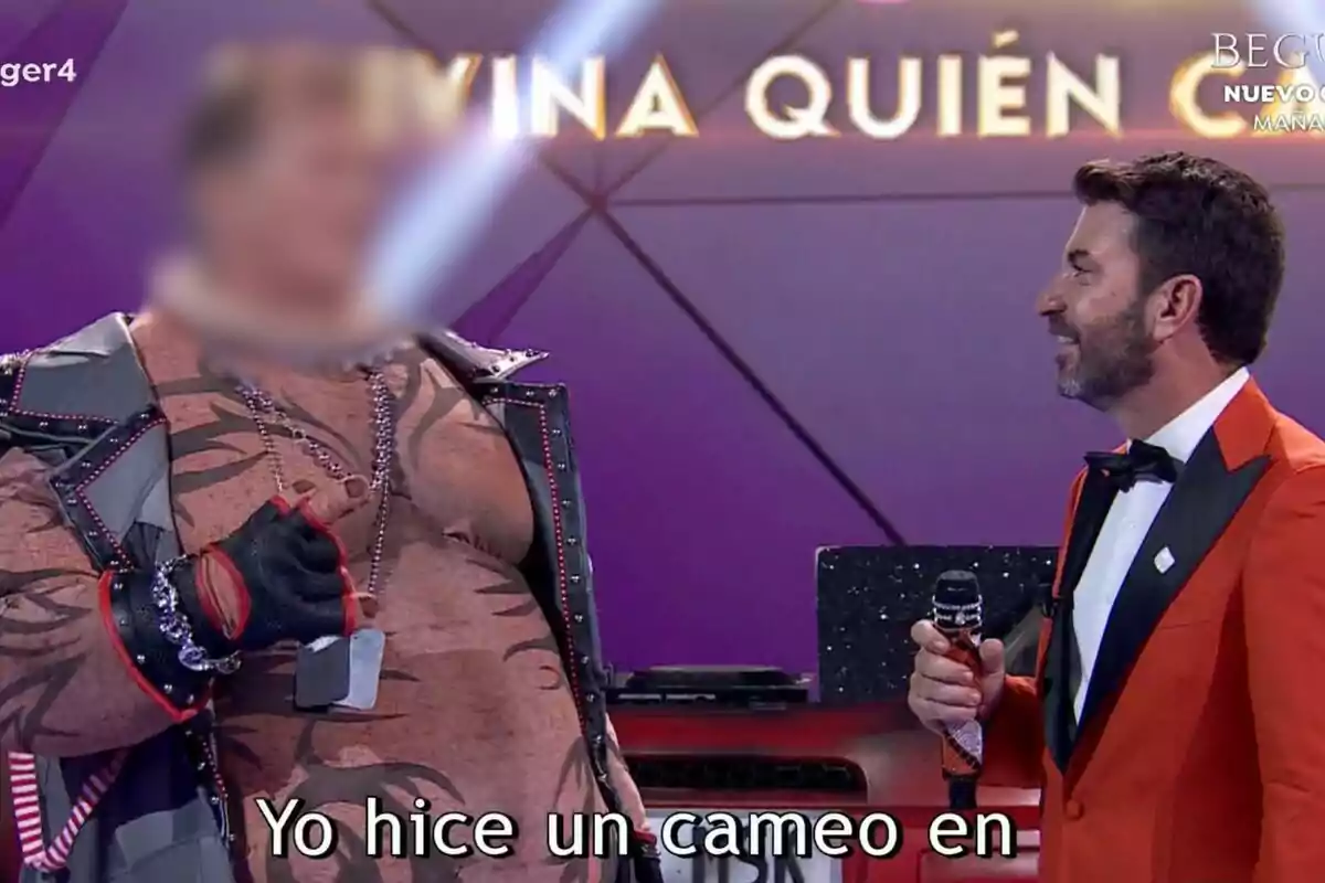 Arturo Valls con un traje naranja sostiene un micrófono mientras habla con David Hasselhoff con la cara difuminada disfrazado en un escenario de Mask Singer.