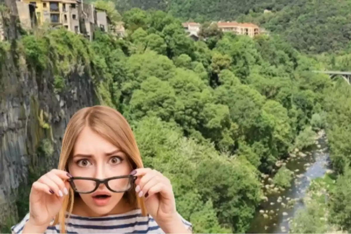 Una mujer con expresión de sorpresa sostiene sus gafas frente a un paisaje de colinas verdes y un río, con casas en el fondo.