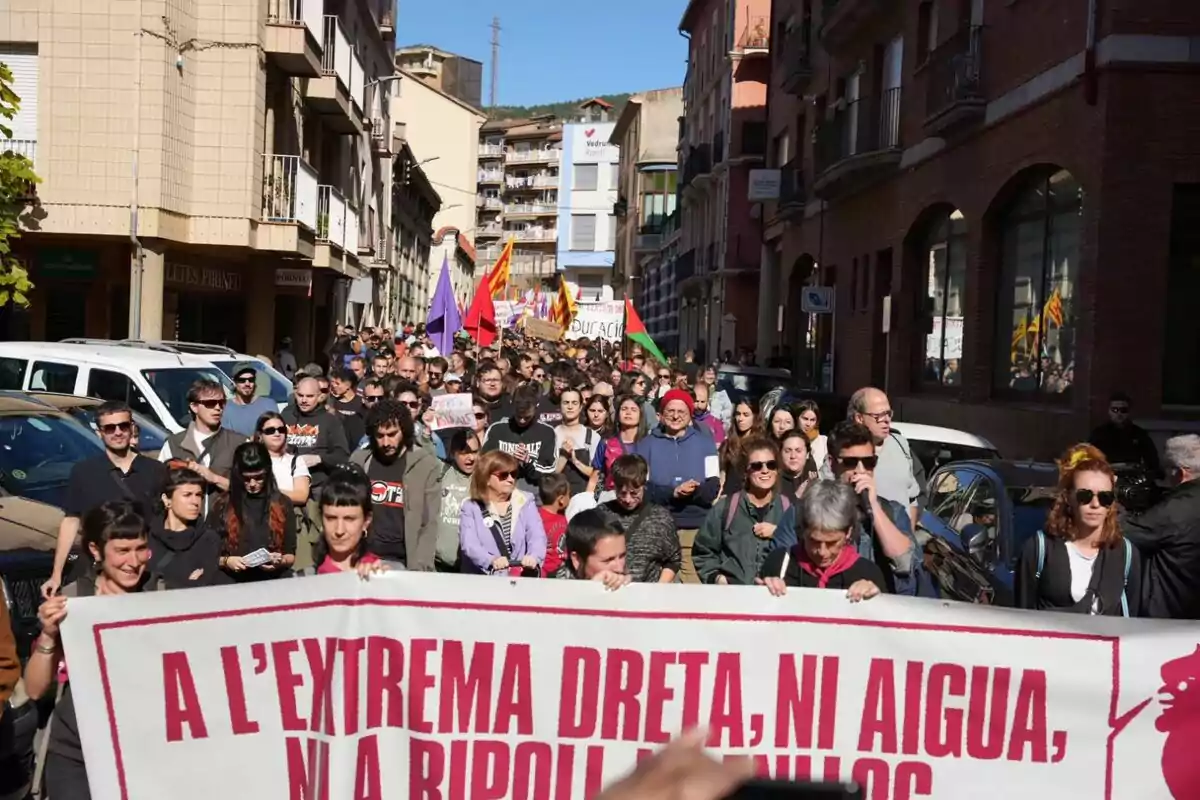 Un grupo de personas participa en una manifestación en una calle, sosteniendo una pancarta con un mensaje en catalán, rodeados de edificios y bajo un cielo despejado.