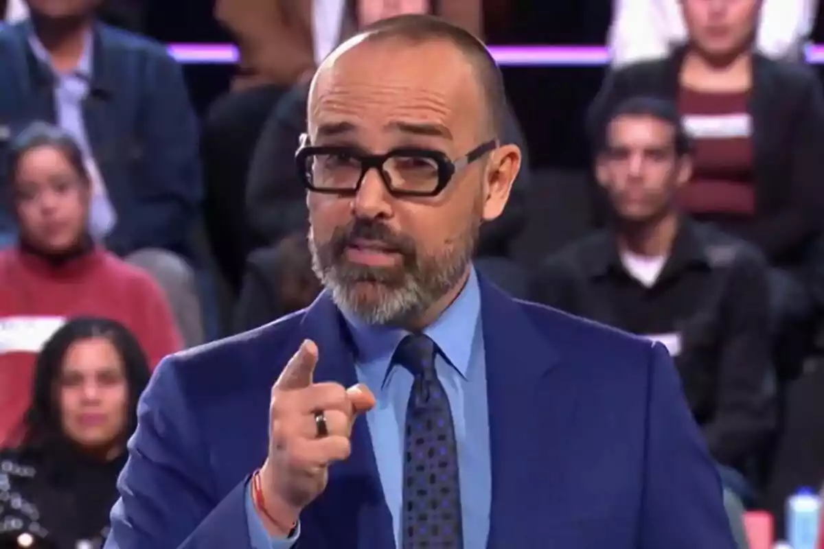 Risto Mejide con gafas y barba vestido con traje azul señala hacia adelante en el plató de Demos: El Gran Sondeo con personas sentadas al fondo.