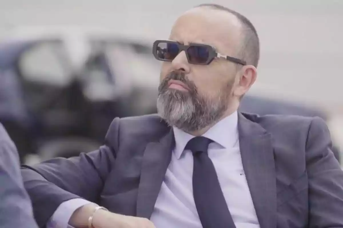 Risto Mejide con barba y gafas de sol vistiendo traje oscuro y corbata sentado al aire libre en 'Viajando con Chester'.