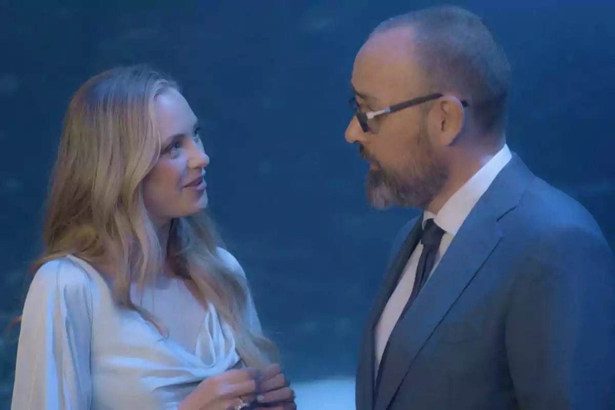 Laia Grassi y Risto Mejide conversan en un ambiente iluminado con luz azul, ella lleva un vestido claro y él viste un traje formal con gafas en Viajando con Chester.