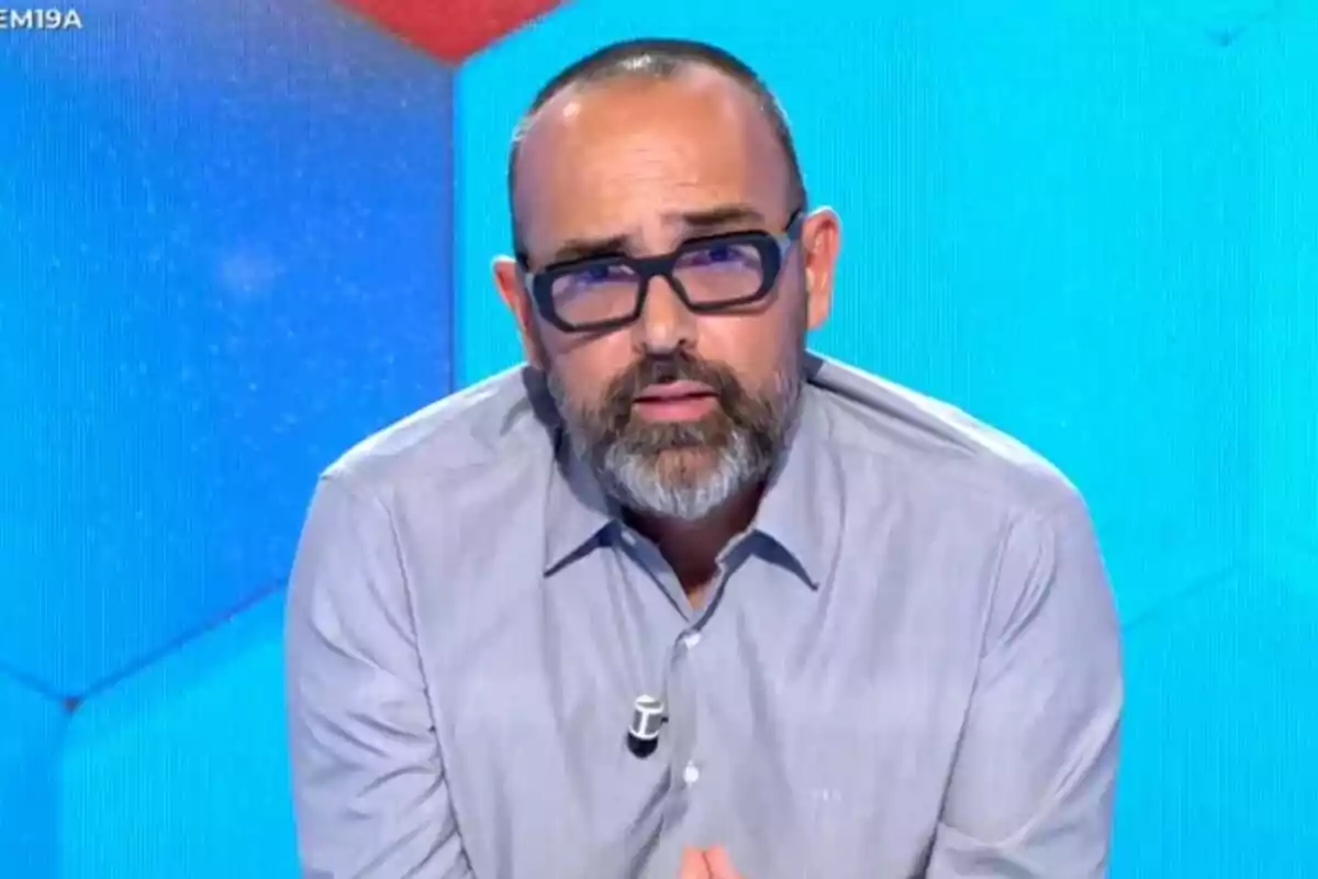 Captura de Risto Mejide con gafas y barba está sentado frente a una pantalla azul y roja, vistiendo una camisa gris y un micrófono de solapa en Todo es Mentira.