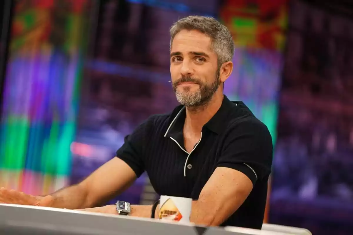 Roberto Leal de cabello corto y barba sentado en una mesa de El Hormiguero con fondo colorido y taza blanca Roberto Leal de cabello corto y barba sentado en una mesa de El Hormiguero con fondo colorido y taza blanca