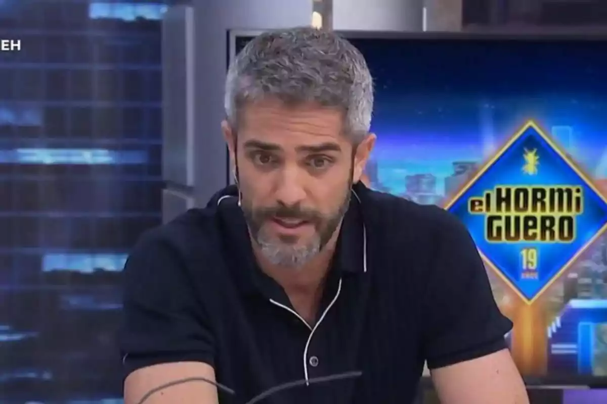 Roberto Leal de cabello canoso y barba hablando en un programa de televisión con el logo de El Hormiguero al fondo