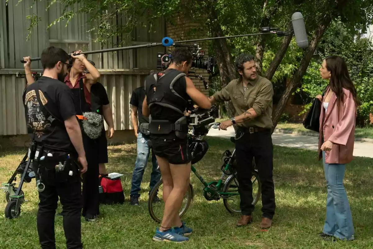 Equipo de filmación grabando una escena al aire libre con dos actores dialogando mientras el equipo técnico sostiene una cámara y un micrófono de boom en la nueva serie comedia 'Sin Gluten' de RTVE. Equipo de filmación grabando una escena al aire libre con dos actores dialogando mientras el equipo técnico sostiene una cámara y un micrófono de boom en la nueva serie comedia 'Sin Gluten' de RTVE.