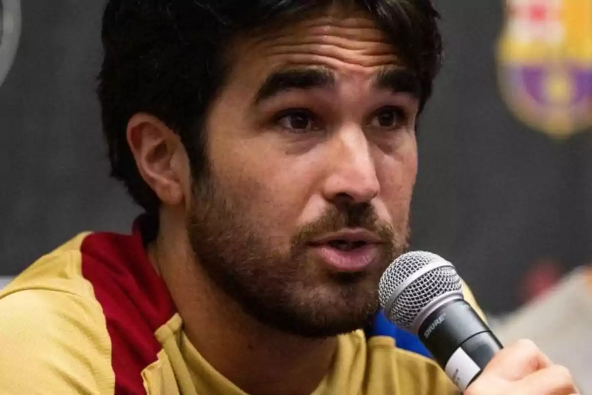 Un hombre con barba y cabello oscuro sostiene un micrófono mientras habla.