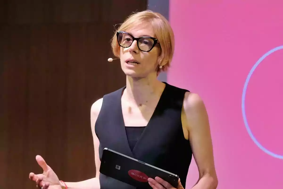 Rosa Romà de cabello corto y rubio con gafas grandes y vestido negro hablando en público mientras sostiene una tableta frente a un fondo rosa de 3Cat