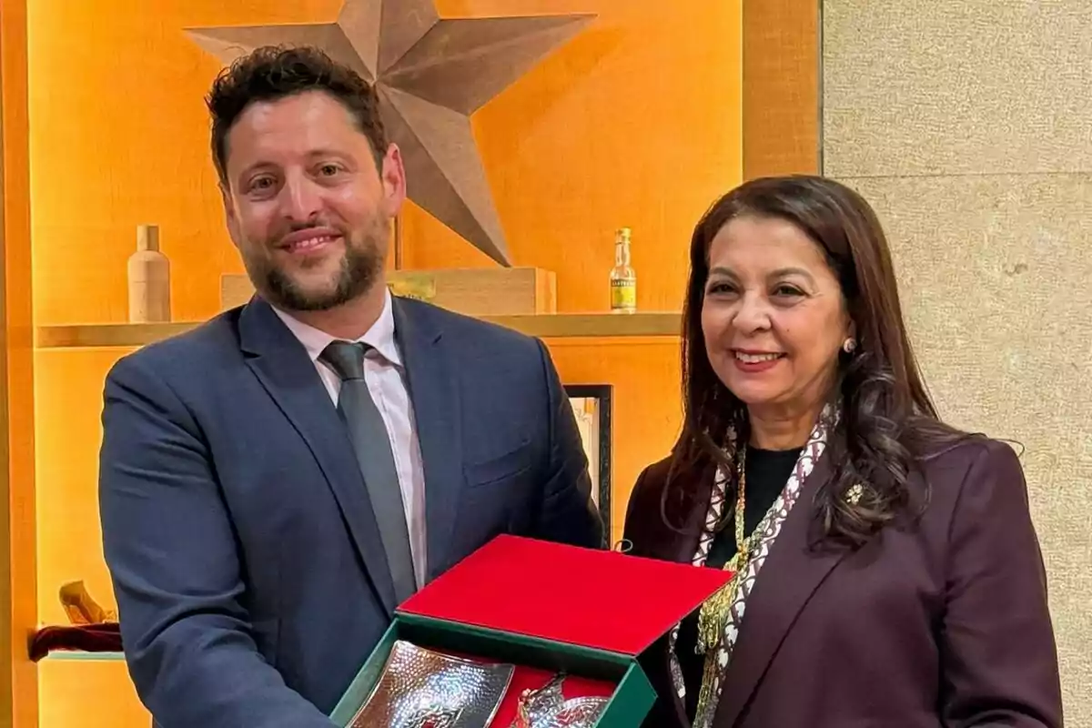 Dos personas posan sonriendo mientras sostienen una caja roja en un entorno interior con una estrella decorativa en la pared. Dos personas posan sonriendo mientras sostienen una caja roja en un entorno interior con una estrella decorativa en la pared.