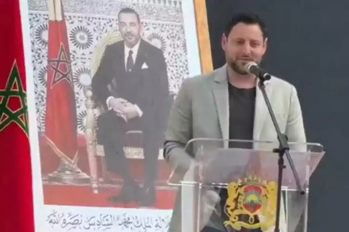 Un hombre hablando en un podio con un micrófono, junto a un retrato de una figura oficial y una bandera con un símbolo de estrella.