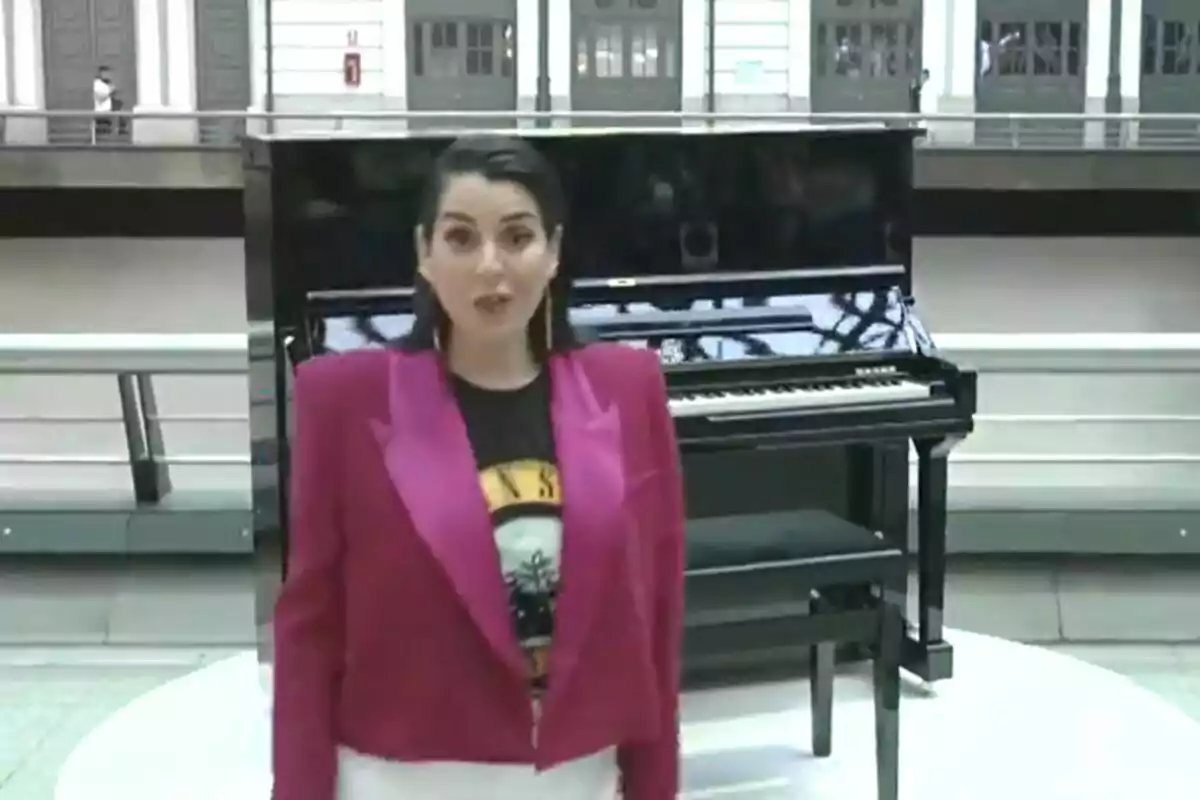 Captura de Ruth Lorezno con chaqueta fucsia y camiseta negra está de pie frente a un piano negro en un espacio interior amplio.