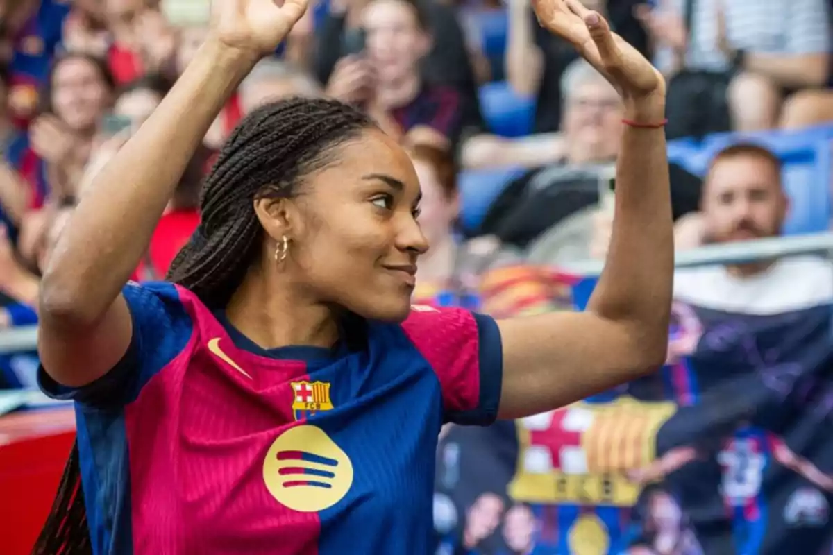 Una jugadora de fútbol con la camiseta del FC Barcelona levanta los brazos mientras sonríe en un estadio lleno de espectadores. Una jugadora de fútbol con la camiseta del FC Barcelona levanta los brazos mientras sonríe en un estadio lleno de espectadores.