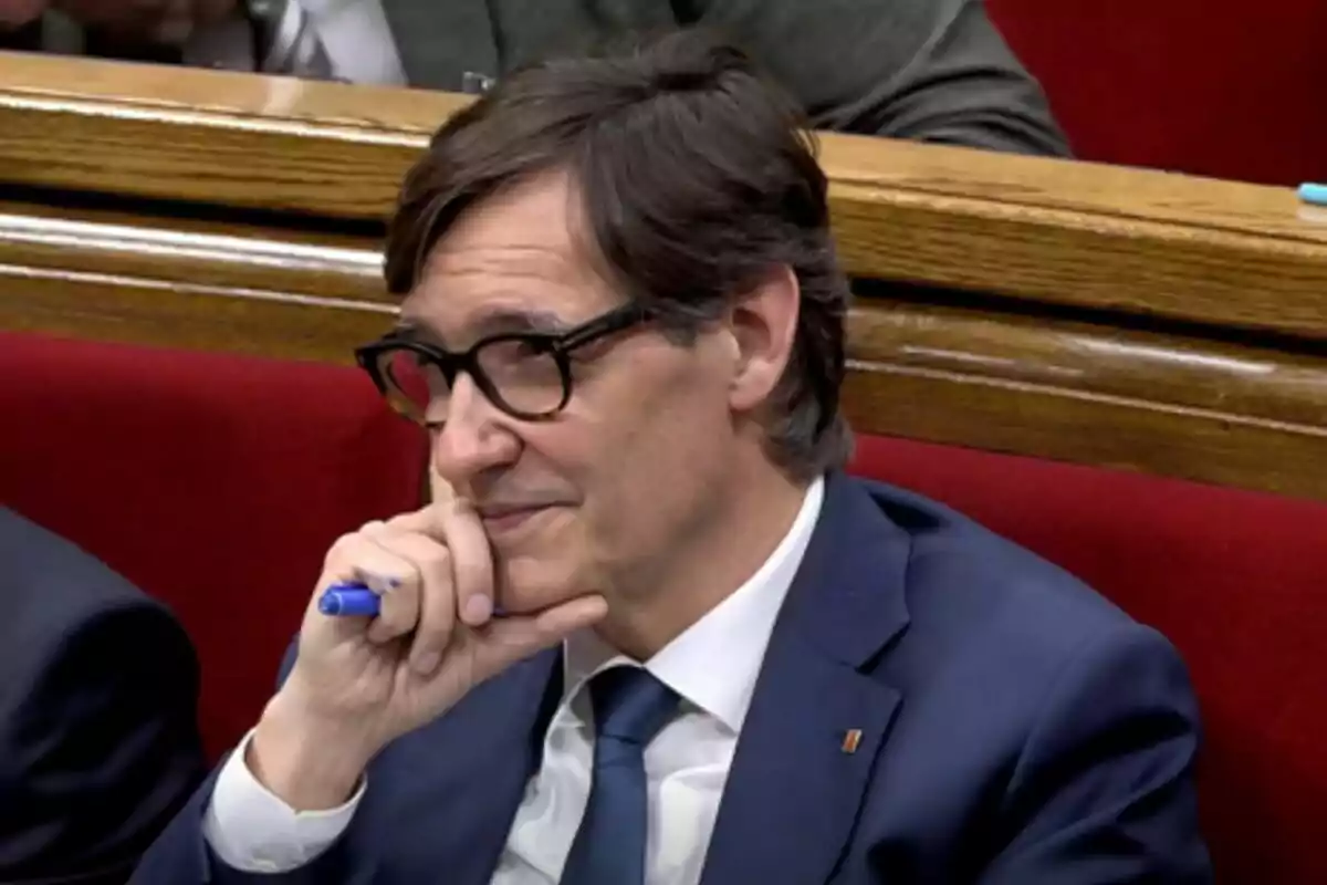 Un hombre con gafas y traje azul sostiene un bolígrafo azul mientras está sentado en un asiento rojo.