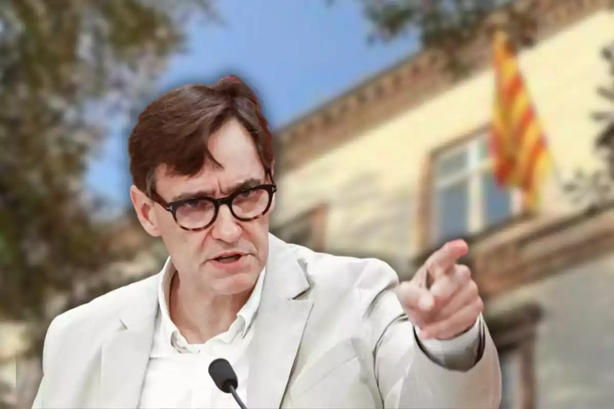 Un hombre con gafas y chaqueta clara gesticula mientras habla frente a un edificio con una bandera catalana.