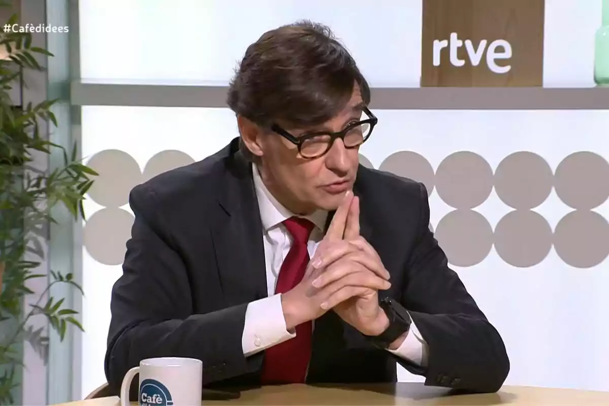 Un hombre con gafas y traje oscuro está sentado en una mesa, con las manos entrelazadas, frente a una taza que dice "Café" y un fondo con el logo de rtve.