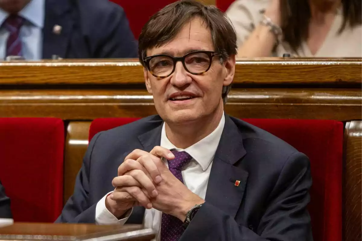 Un hombre con gafas y traje oscuro está sentado en un banco de madera con tapicería roja.