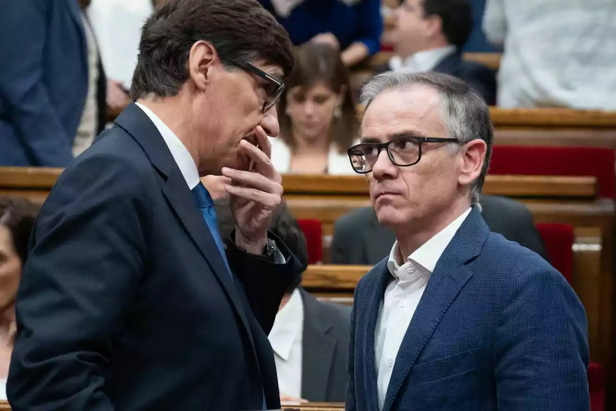 Dos hombres conversando en un entorno formal con personas al fondo.