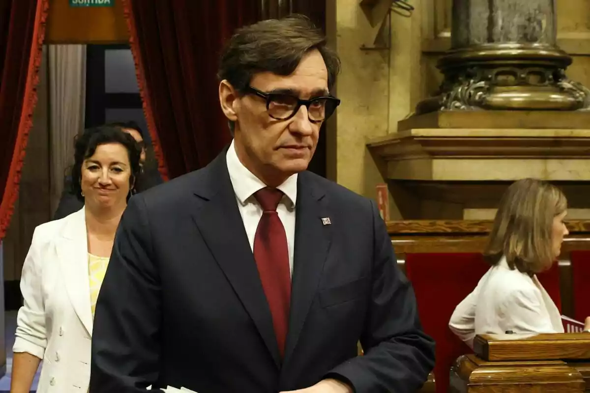 Salvador Illa con traje oscuro con corbata roja está de pie en una sala con cortinas rojas y detalles arquitectónicos clásicos, mientras Alícia Romero con chaqueta blanca sonríe en el fondo.